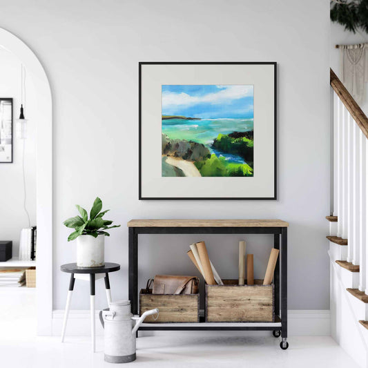 Framed artwork of a coastal scene above a slim table in a white home interior.