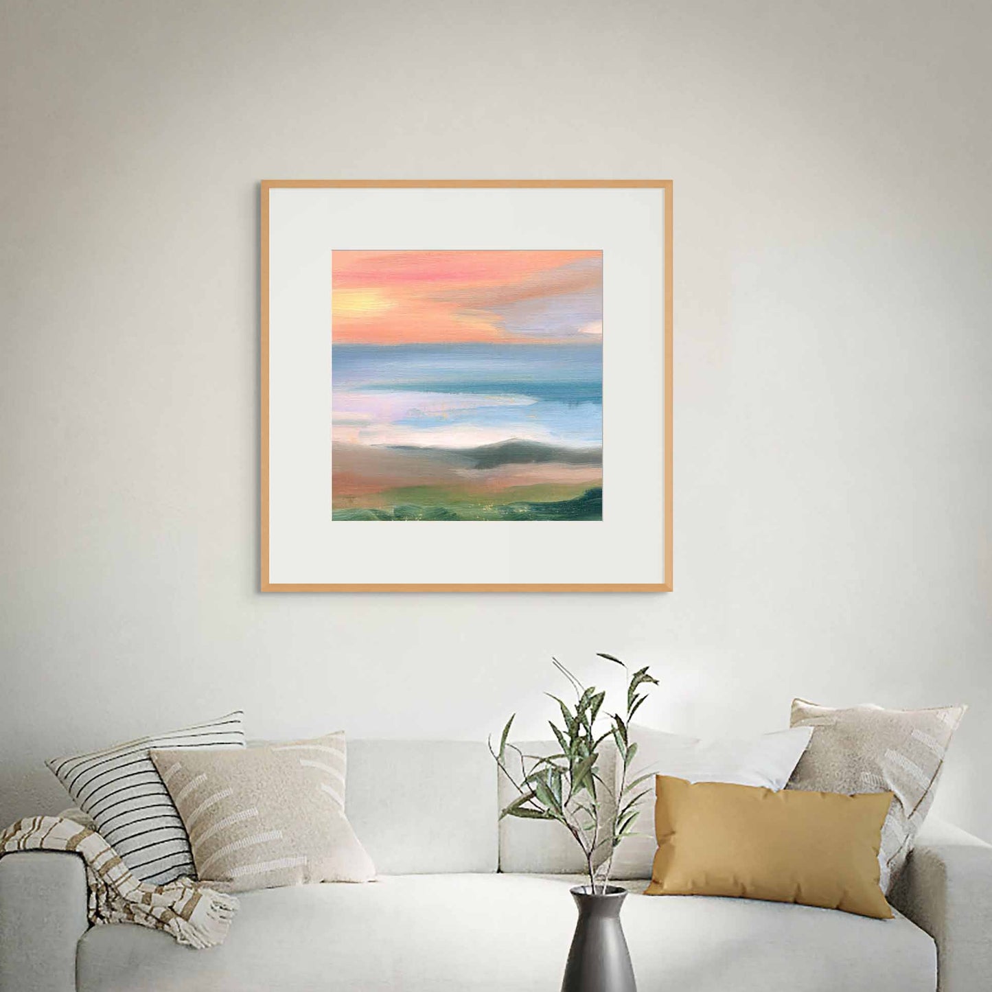 Framed coastal landscape art with loose brush strokes on a wall above a white sofa with cushions and a plant.