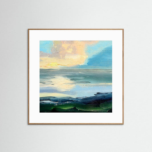Framed artwork of a landscape with a blue sky and orange cloud above the ocean with green stones on a white wall.