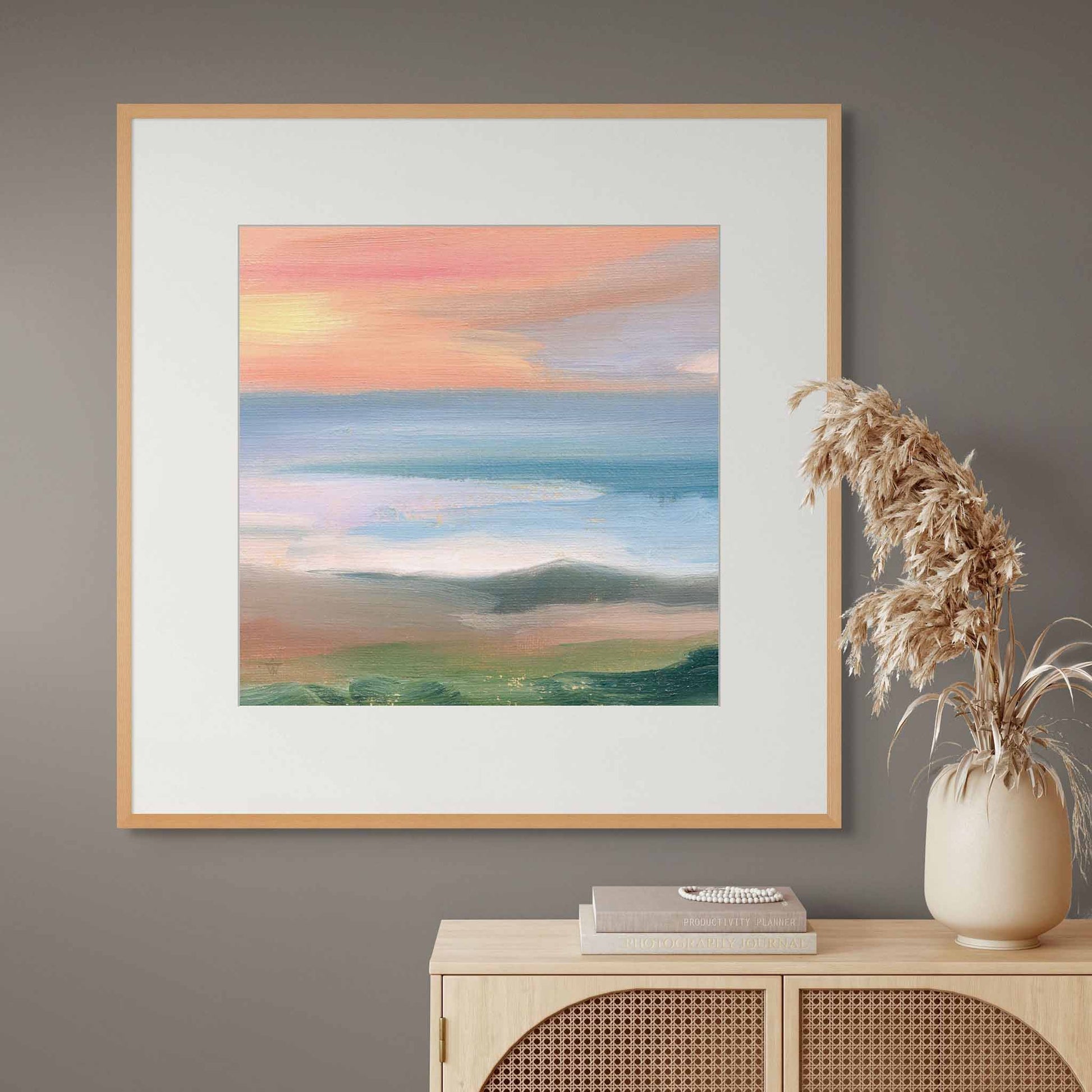 Framed artwork of a landscape with a sunrise over water, displayed on a grey wall above a wooden cabinet.