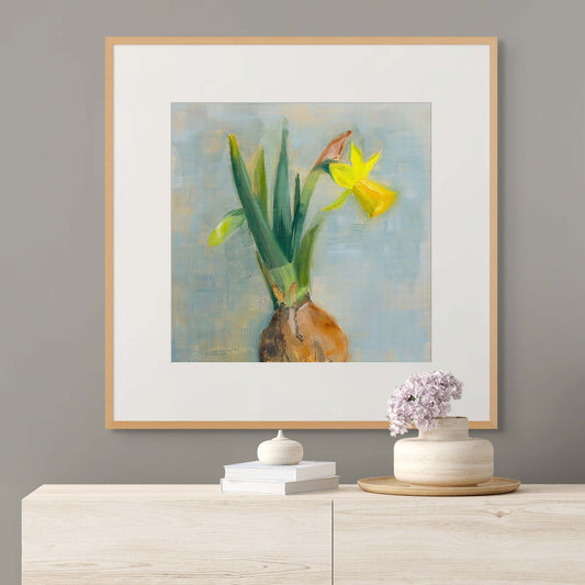 Framed painting of daffodil flowers with bulb on a wall above a wooden cabinet with decorative items.
