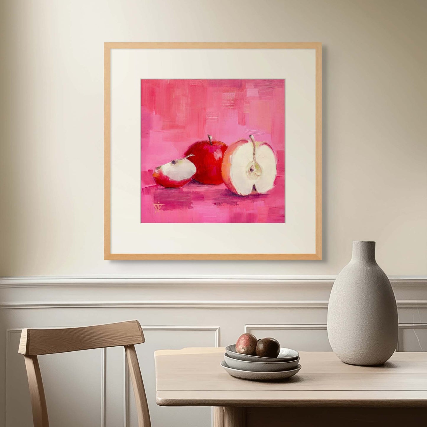 Framed artwork of apples on a pink background above a table in a sunlit dining room setting.