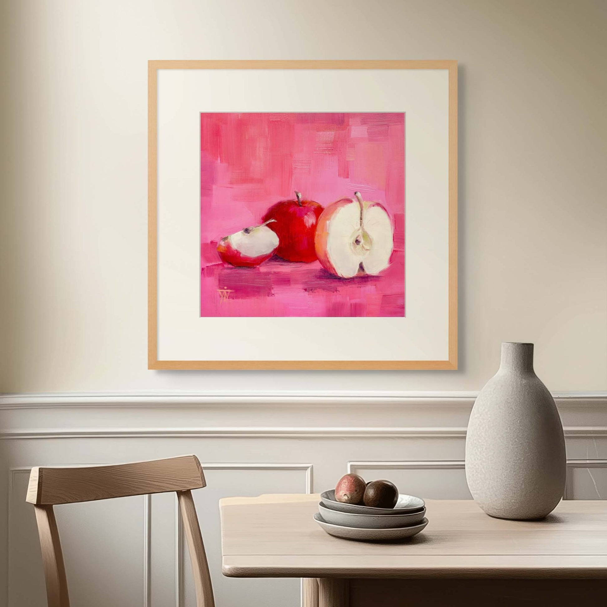 Framed artwork of apples on a pink background above a table in a sunlit dining room setting.