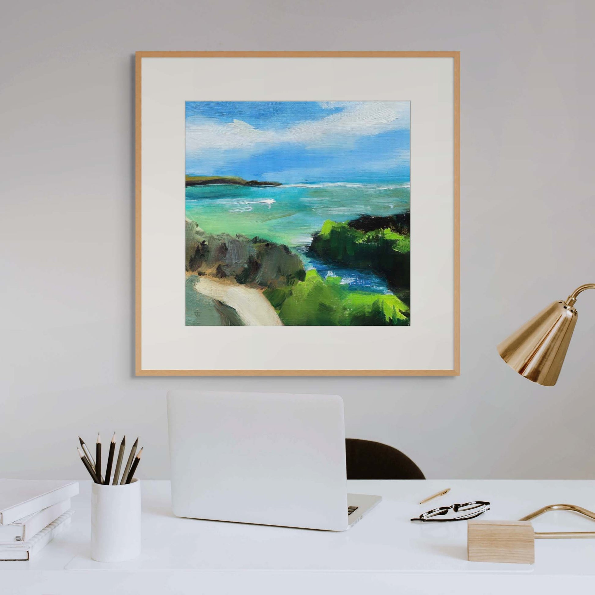 Framed artwork of a coastal scene on a white wall above a white desk with brass lamp and in home office interior.