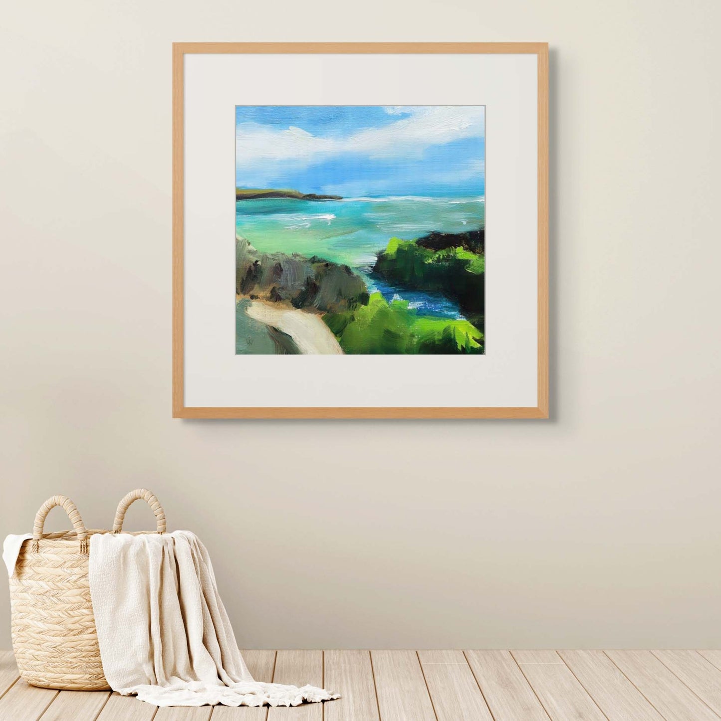 Framed painting of a coastal scene on a wall with a basket and blanket below.
